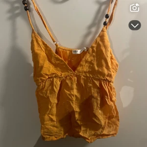 Orangelinnetopp med pärldetaljer - Snygg gul linnetopp med smala axelband dekorerade med svarta pärlor. Toppen har v-ringning och lätt rynkad midja för en avslappnad look. Perfekt för varma dagar och sommarens alla äventyr.