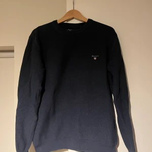 Mörkblå stickad tröja från GANT - Snygg mörkblå stickad tröja från GANT med logga broderad på bröstet. Tröjan har rund hals, ribbade muddar och långärmad passform. Materialet är mjukt och strukturerat, perfekt för kyliga dagar. Passar dig som gillar klassisk och stilren look.