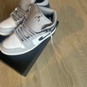 Nike Air Jordan 1 Mid grå/blå - Säljer ett par Nike Air Jordan 1 Mid i grått, vitt och marinblått. Klassisk high-top siluett med snörning, perforerad tå och ikoniska Air Jordan-loggan på sidan. Ovandel i skinn och syntet, perfekt för dig som gillar streetstyle och sneakers med attityd.