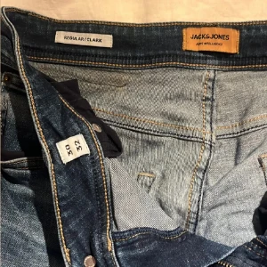 Jack & Jones Regular Clark jeans blå - Säljer ett par Jack & Jones Regular Clark jeans i klassisk blå tvätt med snygga slitningar. Modellen har rak passform, fem fickor och tydliga kontrastsömmar. Jeansen är tillverkade i bomull och har en bekväm midja med knapp och dragkedja.