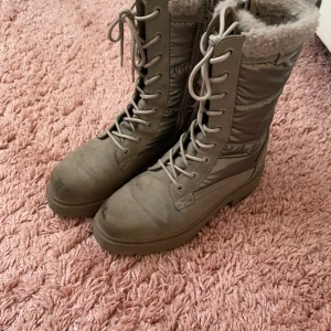 Grå vinterboots med snörning och foder - Säljer ett par höga grå vinterboots med snörning och mjukt, ljust foder. Skorna har platt sula och är tillverkade i syntetmaterial och mocka. Perfekta för kalla dagar med sin varma insida och robusta design.
