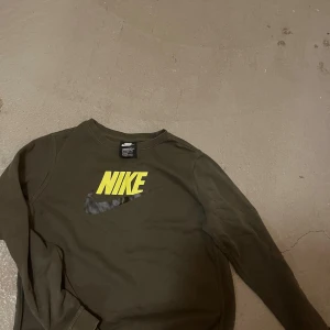 Olivgrön Nike sweatshirt XL - Olivgrön sweatshirt från Nike i bomull med klassiskt gult Nike-tryck och svart swoosh på bröstet. Tröjan har rund hals och långa ärmar, perfekt för en chill och sportig stil.