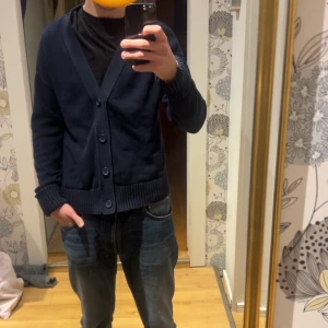 Mörkblå kofta från John Henric - Mörkblå kofta med v-ringning och stora knappar framtill. Tröjan har ribbade muddar vid ärmslut och nederkant, samt en klassisk passform. Perfekt att slänga över en t-shirt för en chill look.