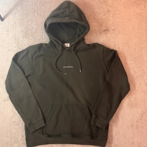 Mörkgrön hoodie från NN07 - Säljer en mörkgrön hoodie från NN07 med broderad text 'NO NATIONALITY' på bröstet. Tröjan har huva med snörning, känguruficka och långa ärmar. Perfekt för dig som gillar stilrena och bekväma plagg.