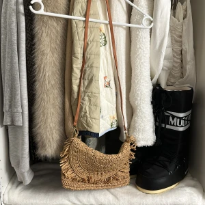Beige axelväska med fransar från H&M - Snygg beige axelväska från H&M i flätad syntet med halvcirkelformad lock och fransdetaljer. Väskan har ett brunt axelband och dragkedja på insidan. Perfekt storlek för mobil, plånbok och småsaker.