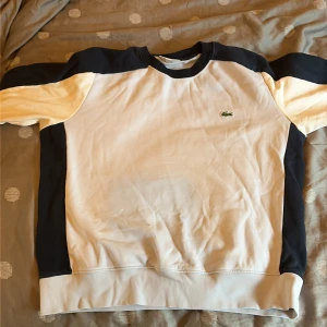 Vit och marinblå sweatshirt Lacoste M - Säljer en stilren sweatshirt från Lacoste i storlek M. Tröjan är vit med marinblå detaljer på axlar, ärmar och sidor. Klassisk rund halsringning och broderad Lacoste-logga på bröstet. Perfekt för en clean och sportig look.