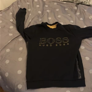 Svart sweatshirt från Hugo Boss S - Svart sweatshirt från Hugo Boss i storlek S. Tröjan har tryckt BOSS-logo framtill och svarta detaljer längs ärmarna med diskret mönster och logga. Klassisk rund halsringning och ribbade muddar. Perfekt för en clean och stilren look.