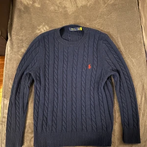 Mörkblå kabelstickad tröja Polo Ralph Lauren - Kabelstickad mörkblå tröja från Polo Ralph Lauren med klassisk röd broderad logga på bröstet. Tröjan har rund halsringning, långa ärmar och ribbade muddar. Perfekt för dig som gillar stilren och tidlös design.