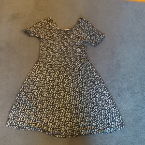 Blommig svart kortklänning - Supersöt svart klänning med vita små blommor över hela tyget. Klänningen har korta ärmar och en lätt utställd kjol. Den har även en stickad överdel.