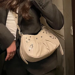 Beige axelväska med nitar och dragkedja - Snygg beige axelväska i skinn med coola silverfärgade nitar och detaljer. Väskan har en bred axelrem med dekorativa sömmar och dragkedja upptill. Formen är rundad och väskan har flera metalldetaljer som ger en edgy vibe.