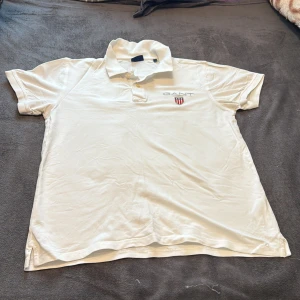 Vit piké från GANT i storlek L - Vit pikétröja från GANT med klassisk krage och korta ärmar. På bröstet finns broderad GANT-logga och ett snyggt emblem med stjärnor och ränder i rött, vitt och blått. Tröjan har knappar vid halsen och är gjord i mjuk bomull som känns skön mot huden.