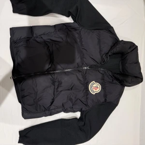 Svart Moncler pufferjacka M - Svart pufferjacka från Moncler med ikonisk logga på bröstet. Jackan har stickade svarta ärmar, huva och dragkedja framtill. Materialet är en mix av dun och stickad polyester, vilket gör den både varm och stilren. Perfekt för dig som vill ha en snygg jacka med streetkänsla.