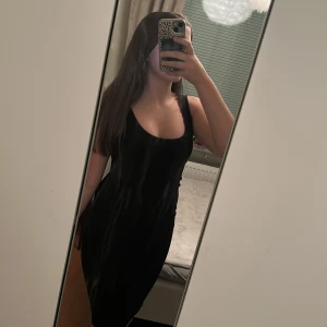 Svart ärmlös klänning - Snygg svart klänning i satin med smala axelband och rundad urringning. Modellen är kroppsnära och slutar ovanför knäna, perfekt för dig som gillar en stilren och enkel look. Passar dig som vill ha en klassisk och tidlös vibe.