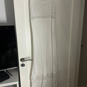 Vit långklänning med spetsdetaljer - Superfin vit långklänning med tunna axelband och vackra spetsdetaljer upptill och nertill. Klänningen har en flowy passform och är tillverkad i ett lätt, skönt tyg som ger en luftig känsla. Perfekt för sommarens varma dagar eller speciella tillfällen. Tyvärr har den blivit för kort på mig.