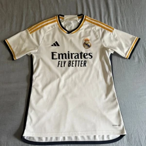 Real Madrid fotbollströja Adidas vit - Snygg Real Madrid fotbollströja från Adidas i vit med svarta och guldiga detaljer. Klassisk design med klubbmärke och sponsortryck på bröstet, samt tre guldiga ränder på axlarna. Tillverkad i lätt och ventilerande material för maximal komfort.