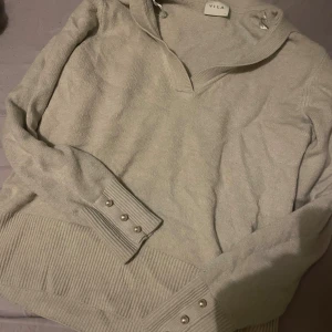 Beige stickad hoodie från Vila - Mysig beige stickad hoodie från Vila med v-ringning och ribbade muddar. Tröjan har långa ärmar med dekorativa pärlknappar vid ärmsluten och en huva med snörning. Perfekt för höst och vinter när du vill vara både bekväm och snygg.