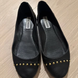 Svarta ballerinaskor Steve Madden - Svarta ballerinaskor från Steve Madden med guldfärgade nitar längs fram- och bakkant. Skorna har rund tå och platt sula. Ovandel i syntetmaterial, innersula i skinn och yttersula i gummi. Snygg och edgy detalj med nitar som ger extra attityd.
