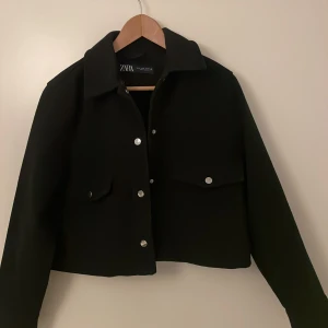 Svart kort kappa från Zara - Stilren svart kort kappa från Zara. Jackan har klassisk krage, stora fickor framtill med lock och silvriga knappar. Perfekt för lager-på-lager och ger en clean look till alla outfits. Andvänt fåtal gånger men ser inga tecken på slitenhet ( passar även S) 