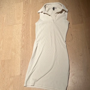 Vit ribbad klänning från H&M XS - Vit ribbad kortklänning från H&M i storlek XS. Klänningen är ärmlös med krage och knappar framtill, vilket ger en sportig och fräsch look. Materialet är mjukt och stretchigt, perfekt för varma dagar.