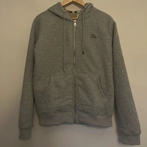 Burberry kofta - Grå hoodie inspirerad av Burberry med dragkedja och klassisk huva. Insidan av luvan har det ikoniska rutmönstret i beige, svart, vitt och rött. Tröjan har snörning i huvan, två fickor fram och ribbade muddar. Perfekt för en chill och stilren look. Tröjan har storlek L men sitter som storlek S