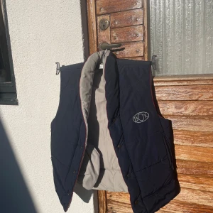 Mörkblå dunväst från BOX - Snygg mörkblå dunväst från BOX med ljusgrå insida och röda detaljer längs kanterna. Västen har broderad logga på bröstet och praktiska fickor med dragkedja. Perfekt för lager-på-lager-stil under kyliga dagar.