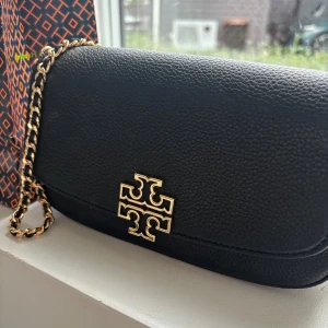 Svart axelväska från Tory Burch - Stilren svart axelväska från Tory Burch i äkta skinn med guldfärgad kedja och ikonisk logga framtill. Väskan har en klassisk, rundad form och magnetlås. Perfekt storlek för det viktigaste och snygg att bära över axeln.