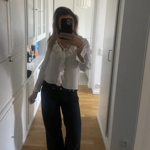 Vit blus med volang och knyt - Säljer en vit blus med långa ärmar, volangdetaljer längs ringningen och framtill samt knytband vid halsen. Blusen har en luftig och lätt känsla, perfekt för en trendig och feminin look.