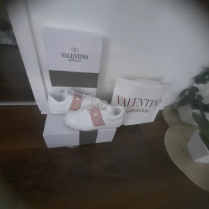 Valentino garavani - Säljer ett par vita Valentino garavani Rockstud sneakers med ikoniska ljusrosa band och nitar över vristen. Skorna har rund tå, platt sula och är tillverkade i skinn. Snygg och lyxig design som sticker ut med sina detaljer och branding på plösen.