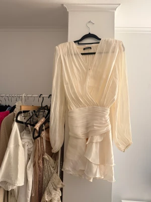 Champagnefärgad omlottklänning med volanger - Superfin kort klänning i champagnefärgat, glansigt material från Ginatricot. V-ringning, långa ärmar och markerad midja. Kjolen har flera lager volanger som ger en flowy känsla. 