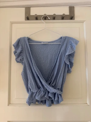 Blå omlottopp med volang från Monki - Supersöt ljusblå omlottopp från Monki i storlek S. Toppen har korta volangärmar, v-ringning och omlottdesign med volangdetaljer längs nederkanten. Materialet är krinklat. Använd ett fåtal gånger. Modellen är ganska kort