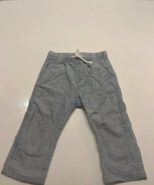 Randiga blå linnebyxor från H&M - Säljer ett par blå och vita randiga linnebyxor från H&M i storlek 80. Byxorna har snörning i midjan, raka ben och knappdetaljer på sidorna samt en ficka bak. Perfekta för chill dagar och sommarhäng.