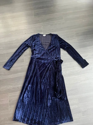 Mörkblå omlottklänning i sammet MW - Snygg långärmad omlottklänning i storlek L men känns som M från Make Way i mörkblå sammet med subtilt mönster. Klänningen har v-ringning, knyt i midjan och ett elegant fall. Perfekt för dig som vill sticka ut med en lyxig känsla och cool textur.