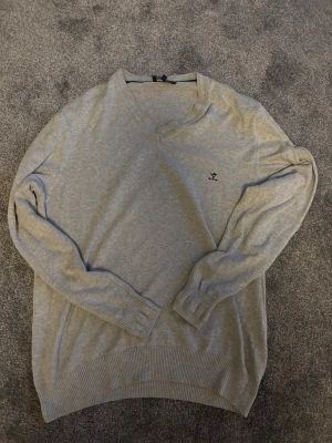 Grå v-ringad tröja Sir Raymond Tailor - Grå v-ringad tröja från Sir Raymond Tailor i storlek L. Tröjan har svarta armbågslappar och ett litet broderat golfmotiv på bröstet. Mjuk och skön kvalitet med ribbade muddar vid ärmslut och nederkant. Perfekt för dig som gillar stilren och sportig look.