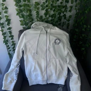 Vit hoodie från Chrome Hearts - Säljer en vit hoodie från Chrome Hearts med svart tryck på bröstet och ryggen. Tröjan har dragkedja, snörning vid huvan och detaljerade mönster längs ärmarna. Materialet är mjuk bomull och passformen är normal. Perfekt för dig som gillar streetwear och snygga detaljer.