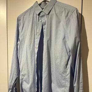 Ljusblå skjorta med knappar - Snygg ljusblå skjorta med klassisk krage och knappar framtill. Skjortan har lång ärm och är tillverkad i mjukt bomullstyg som känns skönt mot huden. Perfekt för dig som gillar stilrena och enkla plagg med clean look.