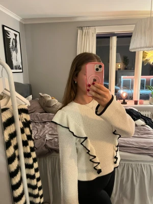 Vit stickad tröja med volangdetaljer - Superfin vit stickad tröja med svarta volangdetaljer längs framsidan och ärmsluten. Tröjan har en rund halsringning och långärmad passform. Den är ribbad vid muddar och nederkant, och har en snygg kontrast mellan vitt och svart.