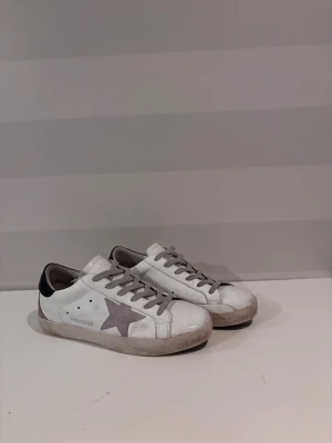 Golden Goose Superstar vita sneakers - Golden Goose Superstar sneakers i vitt skinn med grå mockadetaljer och grå snörning. Sidan har en ikonisk grå stjärna och hälen är svart med logotyp. Platt sula och rund tå ger en klassisk streetstil. Perfekt för dig som gillar en cool och avslappnad look.
