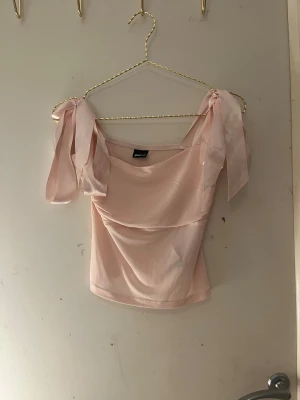 Ljusrosa omlott topp från Gina Tricot - Supersöt ljusrosa topp från Gina Tricot i storlek S. Modellen har omlott-effekt med breda axelband som knyts till rosetter på axlarna. Materialet är mjukt och lätt, perfekt för vår och sommar. Snygg croppad passform som ger en trendig vibe.