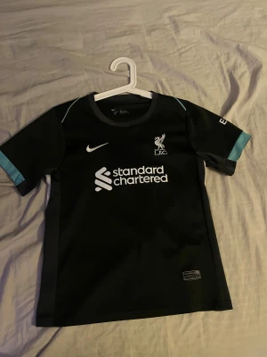 Liverpool FC svart bortatröja Darwin 9 - Snygg svart Liverpool FC bortatröja från Nike med tryck 'Darwin 9' på ryggen. Tröjan har vita och ljusblå detaljer, korta ärmar och klubbmärke samt sponsortryck på bröstet. Perfekt för dig som vill visa ditt stöd för laget och Darwin Núñez.