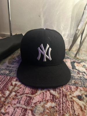Svart New Era Yankees keps 59FIFTY - Säljer en svart New Era 59FIFTY keps med New York Yankees-logga i vitt framtill och broderad frihetsgudinna i blått och vitt på sidan. MLB-logga baktill. Klassisk rak skärm och broderade detaljer. Perfekt för dig som gillar streetwear och sport.