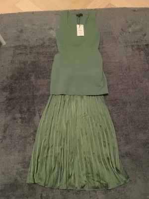 Grön långklänning från Zara - Snygg grön långklänning från Zara med ärmlös överdel och plisserad kjol. Klänningen har en stilren, figurnära passform upptill och ett flowy fall nertill. Perfekt för dig som vill sticka ut med en modern och fräsch look.