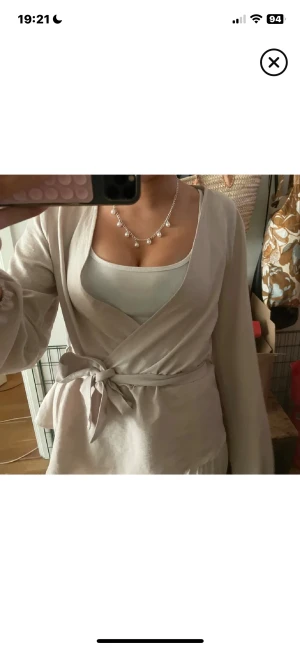 Beige omlottopp med knytband - Säljer en beige omlottopp med långa ärmar och snygg knytning i midjan. Toppen har en v-ringad design och är gjord i mjukt bomullsmaterial. Perfekt för dig som gillar en chill och stilren look med lite extra detalj vid midjan.