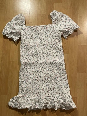 Vit blommig kortklänning från Gina Tricot - Vit kortklänning från Gina Tricot med små blommor. Klänningen har puffiga kortärmar, smockad kropp och volangkant nedtill.