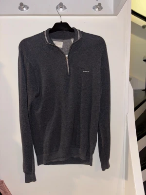 Mörkgrå half zip tröja från GANT - Snygg mörkgrå långärmad tröja från GANT med half zip och diskret logga på bröstet. Tröjan har en klassisk krage med ljusa detaljer och är tillverkad i mjukt stickat material.