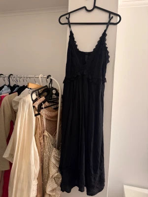 Svart långklänning med spetsdetaljer - Svart långklänning med tunna axelband och v-ringning. Klänningen har fina spetsdetaljer längs urringningen och under bysten samt nertill. 