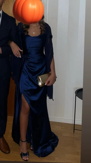 Marinblå långklänning i satin - Elegant marinblå långklänning i glansigt satinliknande material med draperad överdel och offshoulder-detalj. Klänningen har en snygg slits framtill och smala axelband. Perfekt för bal eller festliga tillfällen när du vill sticka ut.