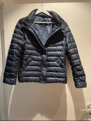 Mörkblå pufferjacka från GANT - Snygg mörkblå pufferjacka från GANT i storlek small. Jackan har horisontella quiltade sektioner, hög krage och dragkedja framtill. Tillverkad i polyester, perfekt för kyliga dagar. Enkel och stilren design som funkar till många olika looks.