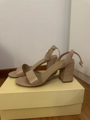 Beige sandaler med blockklack från XIT - Snygga beige sandaler med breda remmar och blockklack. Skorna har krokodilpräglat mönster och justerbart spänne vid ankeln. Rund tå och öppen häl ger en fräsch vibe, perfekt till sommarens outfits. Materialet är syntet med skinnliknande finish.