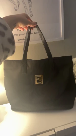 Svart Fendi vintage shoppingväska - Snygg svart shoppingväska från Fendi med klassisk guldlogga framtill. Väskan har två rejäla handtag och är tillverkad i skinn med insida i svart tyg. Inuti finns en guldfärgad dragkedja och Fendi-plakett. Perfekt för dig som gillar lyxiga och tidlösa accessoarer.
