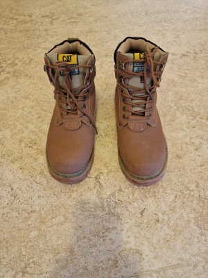 Bruna CAT boots i skinn - Stabila bruna boots från CAT. Skorna är tillverkade i skinn och har snörning framtill samt vadderad kant i svart. Perfekta för dig som gillar robust stil och vill ha något slitstarkt till höst och vinter.